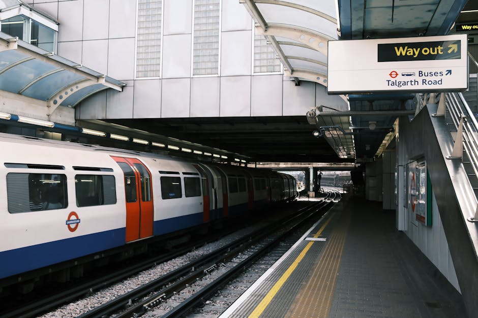 A modern London Underground train with white, blue, and red exterior is stationary at a platform near Ickenham tube station. The platform features a textured, non-slip surface with a yellow tactile st