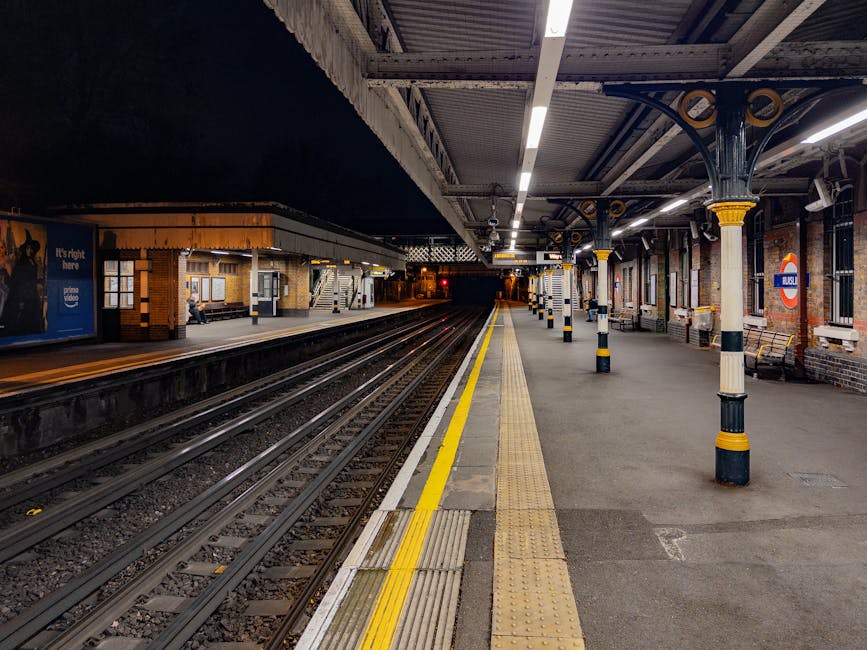 A modern London Underground train with white, blue, and red exterior is stationary at a platform near Ickenham tube station. The platform features a textured, non-slip surface with a yellow tactile st