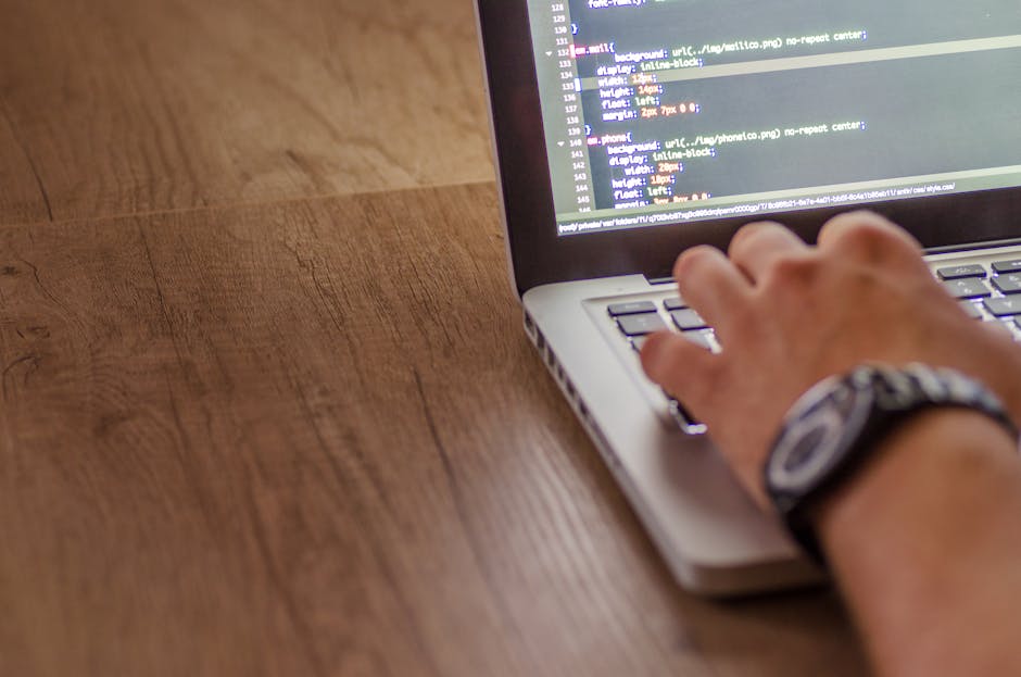 A person's hand, wearing a wristwatch, is seen typing on a laptop keyboard placed on a wooden desk. The laptop screen displays lines of code, suggesting programming or website management activities. T